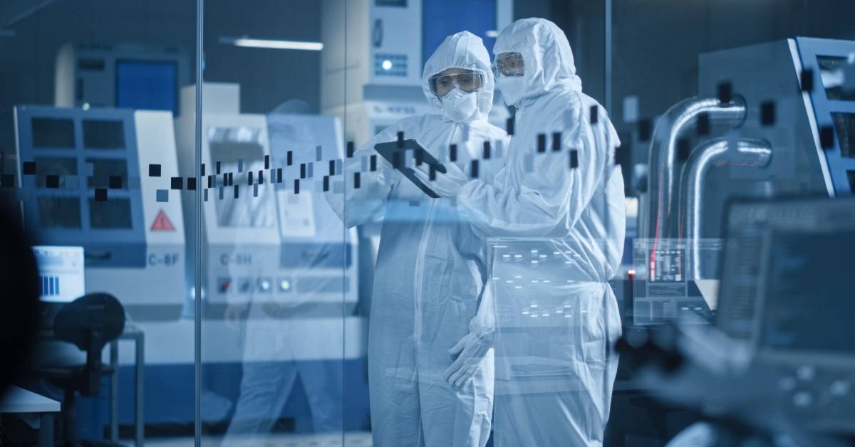 Two employees wearing safety goggles and coveralls. One employee holds a tablet while the other looks at it.