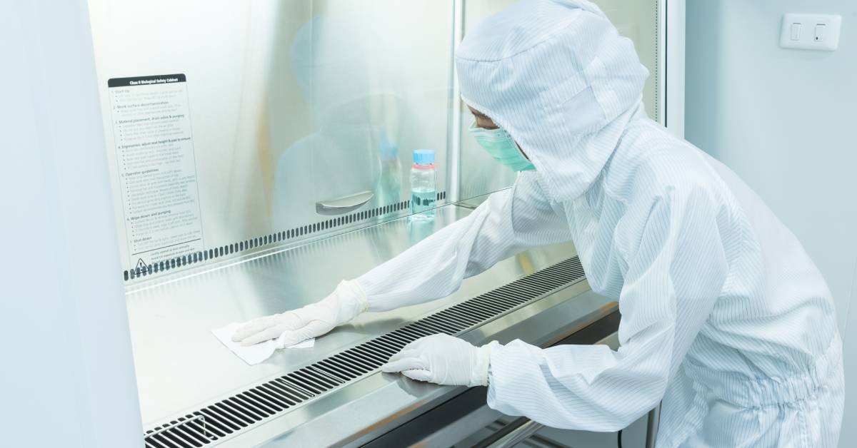 An employee wearing a white coverall, wiping a shiny metal table. The metal table has a bottle with blue liquid on it.