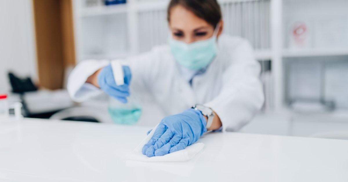 A person wearing a blue face mask and blue gloves wipes a white surface and holds a spray bottle filled with blue liquid.