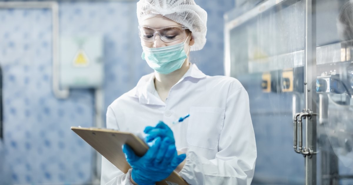 An employee wears a blue surgical mask, a white cap, and blue gloves. They are writing on a clipboard.