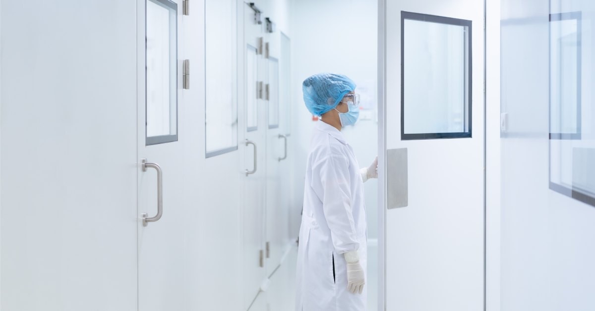 An employee wears a blue cap and mask while opening a door in a hallway. The hallway is full of white walls and doors.