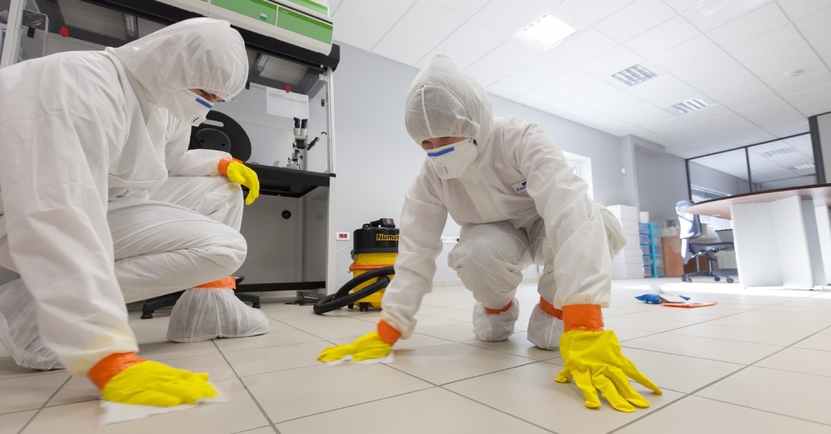 Two employees wearing white lab gowns and yellow gloves are kneeling down on a laboratory floor to wipe it clean.