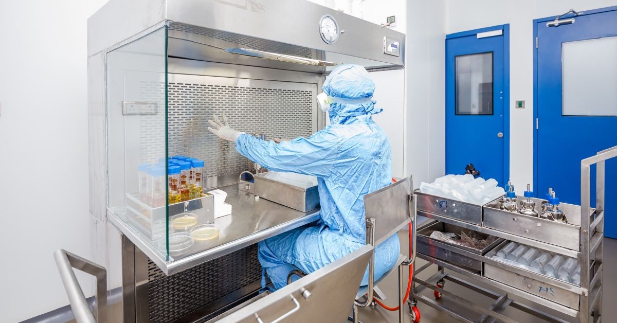 A person wearing a blue lab gown sits in a metal chair. The desk in front of them has vials and petri dishes on it.