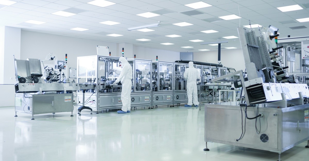 Two employees are dressed head-to-toe in PPE while working in a brightly lit laboratory that is full of metal machines.
