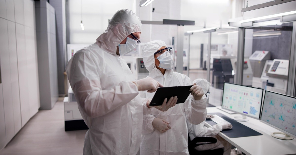 Two employees wearing PPE are holding a tablet. Another employee is sitting and working at a computer nearby.